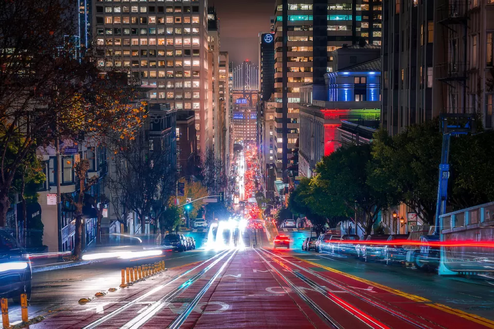 Blurred lights of traffic in motion on San Francisco street with high-rise buildings and Bay Bridge visible in background at night.